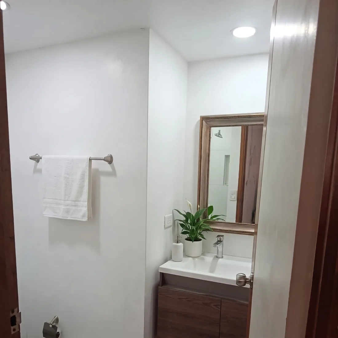 Baño blanco con lavamanos sobre mueble de madera y espejo, con una toalla, una planta y un dispensador agregados virtualmente.