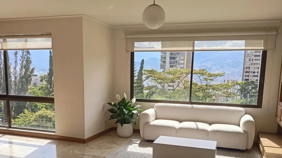 Sala con dos ventanales hacia los árboles y la montaña, con amoblamiento virtual: sofá blanco, mesa baja, alfombra y una planta.